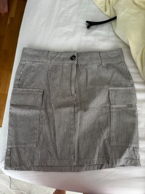 Randig kortkjol från Gina Tricot - Säljer en snygg kortkjol från Gina Tricot med smala vertikala ränder i blått och vitt. Kjolen har två stora fickor framtill och stängs med knapp och dragkedja. Perfekt till sommaren och enkel att matcha med olika toppar.