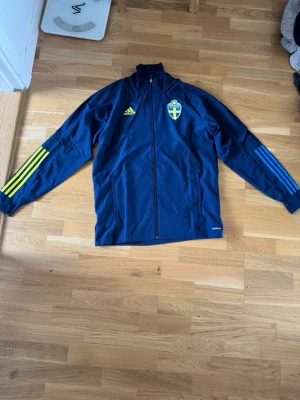 Sverige träningsjacka från Adidas - Mörkblå träningsjacka från Adidas med Sveriges fotbollslandslagsmärke på bröstet. Jackan har hel dragkedja, ståkrage och gula samt blå ränder på ärmarna. Perfekt för dig som vill representera Sverige på träning eller match.