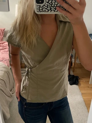 Beige omlottopp med knyt - Snygg beige omlottopp med korta ärmar och knyt i sidan. Toppen har v-ringad hals och en figurnära passform som framhäver midjan. Perfekt att styla med jeans eller kjol för en trendig look.