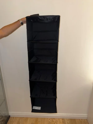 Svart hängande förvaringshylla - Praktisk svart hängande förvaringshylla i tyg med fem fack. Perfekt för garderoben eller hallen för att organisera kläder, skor eller accessoarer. Enkel att hänga upp med kardborreband upptill och tar minimalt med plats när den inte används.