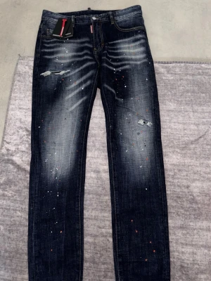 Mörkblå Dsquared2 jeans med slitningar - Säljer ett par mörkblå jeans från Dsquared2 med coola slitningar och färgstänk. Jeansen har en rak passform, klassiska fem fickor och detaljerade slitningar på låren. Materialet är denim med en snygg tvätt och kontrastsömmar. Perfekta för en trendig streetwear-look.