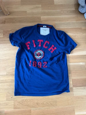 Blå Abercrombie & Fitch t-shirt 2000 stil - Snygg blå t-shirt från Abercrombie & Fitch med röd broderad text 'FITCH 1892' och ett emblem på bröstet. Klassisk rund halsringning och korta ärmar. Perfekt för en avslappnad och trendig stil.