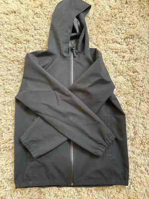Svart regnjacka från Uniqlo - Snygg svart regnjacka från Uniqlo med huva och hel dragkedja framtill. Jackan har två sidofickor och elastiska muddar vid ärmsluten. Tillverkad i ett vattenavvisande material som passar perfekt för regniga dagar.