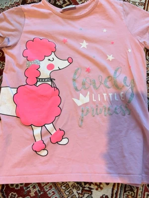 Rosa t-shirt med pudeltryck - Söt rosa t-shirt med tryck av en pudel och texten 'lovely little princess' i glittriga bokstäver. T-shirten har korta ärmar och rund halsringning. Perfekt för dig som gillar gulliga motiv och mjuka material.