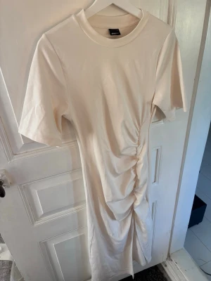 Beige klänning från Gina Tricot - Trendig beige klänning från Gina Tricot med rund halsringning och korta ärmar. Klänningen har snygg rynkning på ena sidan som ger en cool twist till looken. Perfekt för dig som gillar enkel men stilren design. Aldrig använd.
