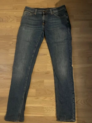 Blå raka jeans från Nudie Jeans - Säljer ett par klassiska blå jeans från Nudie Jeans med rak passform och fem fickor. Jeansen har subtila slitningar och kontrastsömmar, tillverkade i denim av bomull. Perfekta för en avslappnad och stilren look.