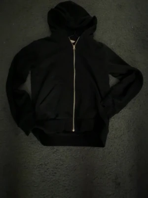 Svart zip-hoodie med huva - En klassisk svart hoodie med dragkedja framtill och huva. Hoodien har två fickor på magen och ribbade muddar vid ärmslut och nederkant. Perfekt för en avslappnad och stilren look.