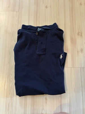 Marinblå pikétröja från Ralph Lauren - Klassisk marinblå pikétröja från Ralph Lauren med korta ärmar och den ikoniska broderade loggan på bröstet. Tröjan har krage, knappar vid halsen och är tillverkad i mjuk bomull. Perfekt för en avslappnad men stilren look.