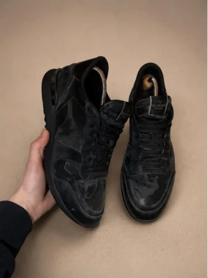 Valentino rockrunners  - Säljer ett par svarta sneakers från Valentino med mörkgrönt camomönster. Skorna har låga skaft, klassisk snörning och är tillverkade i en mix av mocka och textil. Men tyvärr lite hål i hälen efter andvändning. Snygg och stilren design med diskret logga på plösen. Perfekta för dig som gillar exklusiva skor. Pris går att diskutera.