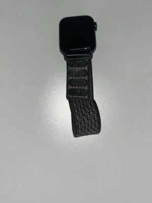 Svart Apple Watch med nylonarmband - Apple Watch SE i svart med ett justerbart svart nylonarmband. Klockan har en fyrkantig form och digital krona på sidan. Klockan har 1 repa över skärmen men annars fungerar den felfritt!!