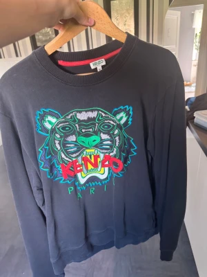 Svart Kenzo sweatshirt med tigertryck - Cool svart sweatshirt från Kenzo med ikoniskt broderat tigerhuvud i grönt, blått och vitt samt röd Kenzo-logga och texten Paris. Tröjan har rund halsringning och ribbade muddar. Perfekt statement-plagg för dig som gillar streetwear.