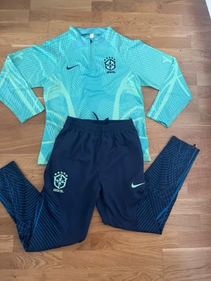 Nike Brasil träningsset blå/turkos - Snyggt träningsset från Nike med Brasil-tryck. Överdel i turkos med vita och ljusgröna mönster, halv dragkedja och lång ärm. Matchande marinblå byxor med turkosa detaljer och elastisk midja. Perfekt för fotbollsträning eller sportiga dagar.