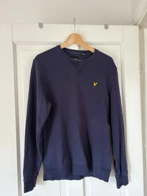 Mörkblå sweatshirt från Lyle & Scott - Mörkblå sweatshirt från Lyle & Scott med klassisk rund halsringning och gul logga broderad på bröstet. Tröjan har ribbade muddar vid ärmslut och nederkant samt en avslappnad passform. Perfekt för en stilren och enkel look.