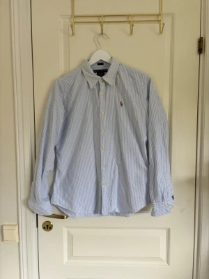 Blårandig skjorta från Ralph Lauren - Klassisk blårandig skjorta från Ralph Lauren med vit knappstängning och broderad logga på bröstet. Skjortan har lång ärm, krage och är tillverkad i bomull. I nyskick.