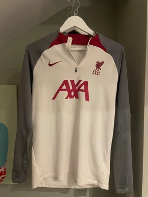 Liverpool Nike långärmad träningströja - Snygg långärmad träningströja från Nike med Liverpool FC-logga och AXA sponsortryck. Tröjan är vit med grå ärmar och röda detaljer vid axlar och tryck. Halv dragkedja framtill och sportig passform, perfekt för träning eller uppvärmning.