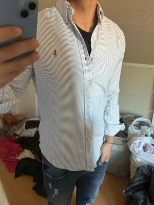 Randig skjorta från Ralph Lauren - Klassisk vit skjorta med tunna blå ränder från Ralph Lauren. Skjortan har lång ärm, knappar framtill och den ikoniska broderade loggan på bröstet. Tillverkad i bomull och har en normal passform. Perfekt för en clean och stilren look.
