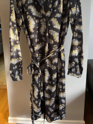 Mönstrad klänning från Lindex - Mörkblå omlottklänning från Holly & Whyte by Lindex med blommigt och paisley-inspirerat mönster i gult, vitt och blått. Klänningen har lång ärm, knytband i midjan och en mjuk, följsam passform. Perfekt för dig som gillar färg och mönster!