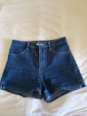 Mörkblå högmidjade jeansshorts - Snygga mörkblå jeansshorts med hög midja och klassisk femficksdesign. Shortsen har bälteshällor, knapp och dragkedja framtill samt raka ben. Perfekta för varma dagar och enkla att matcha med olika toppar.