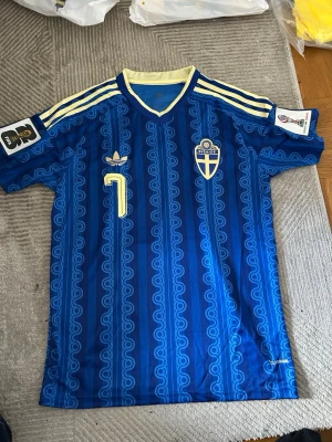 Blå Sverige fotbollströja Adidas - Retro blå fotbollströja från Adidas med Sveriges landslagsemblem på bröstet och nummer 7. Tröjan har gula detaljer, klassiska Adidas-ränder på axlarna och ett unikt mönster i tyget. Perfekt för dig som älskar fotboll och vill sticka ut på planen.
