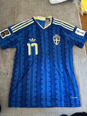 Blå Sverige matchtröja Adidas - Säljer en blå Sverige matchtröja från Adidas med retroinspirerat mönster, gula detaljer och nummer 17 på bröstet. Tröjan har broderat landslagsemblem, Adidas-logga och tre gula ränder på axlarna. Tillverkad i lätt och ventilerande syntetmaterial.