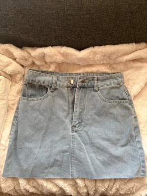 Ljusblå jeanskjol med fickor - Säljer en ljusblå jeanskjol i klassisk modell med fickor, bälteshällor och dragkedja framtill. Kjolen har en rak passform och är tillverkad i denim.