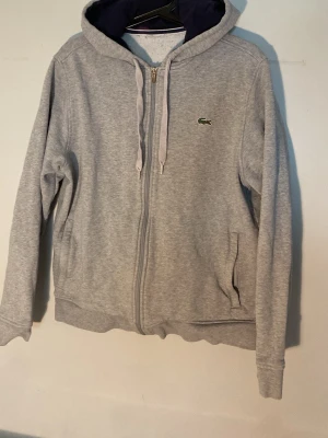 Grå zip-hoodie från Lacoste - Snygg grå hoodie från Lacoste med dragkedja och klassisk krokodil-logga på bröstet. Mjuk bomullsmix, två fickor fram och justerbar huva med snören. Perfekt för en chill och sportig look.