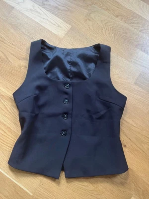 Svart figursydd väst med knappar - Stilren svart väst med fyra knappar framtill och figurnära passform. Västen har djup urringning och är helfodrad med slät insida. Perfekt att styla över en skjorta eller t-shirt för en trendig look.