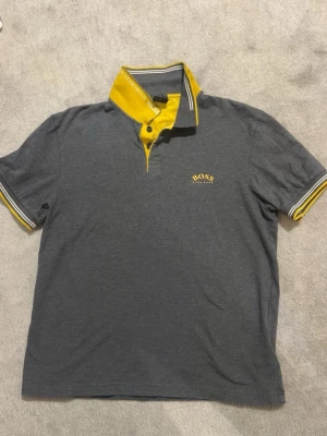 Hugo Boss pikétröja grå/gul strl M - Säljer en Hugo Boss pikétröja i mörkgrå färg med gula detaljer och broderad Boss-logga på bröstet. Stilren och bekväm modell i fint skick utan hål eller fläckar. Storlek M (passar även S beroende på passform) ✅ Mycket fint skick ✅ Original Hugo Boss ✅ Broderad logga