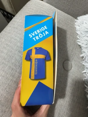 Sverige supporter t-shirt blå/gul - Supporter t-shirt i blått och gult med ett stort kors i gult och texten SWEDEN på framsidan. Perfekt för att visa ditt stöd för Sverige. Klassisk passform och korta ärmar.