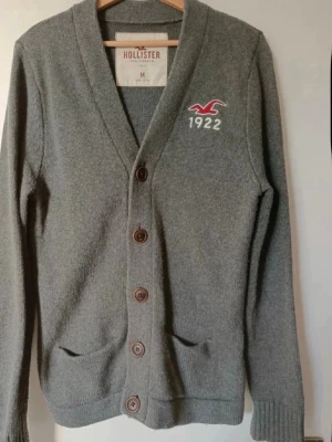 Grå kofta från Hollister med knappar - Mysig grå kofta från Hollister med knäppning framtill och två fickor. Broderad logga och siffran 1922 på bröstet. Perfekt för lager-på-lager och har en klassisk v-ringad design.