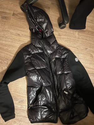 Svart pufferjacka med huva från Moncler - Snygg svart pufferjacka från Moncler med glansig yta och huva. Jackan har svarta tygärmar, två dragkedjeförsedda fickor och Moncler-logga på ärmen. Perfekt för kalla vinterdagar och riktigt trendig look.