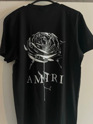 Svart Amiri t-shirt med tryck - Säljer en svart t-shirt från Amiri i mjuk bomull. Framsidan har ett coolt tryck med Amiri-loggan och färgstänk, medan baksidan pryds av en stor vit ros och Amiri-tryck. Klassisk passform och rund hals. Perfekt för dig som gillar streetwear och snygga detaljer.