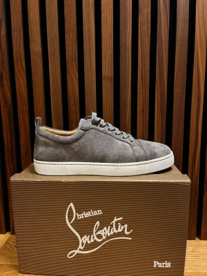 Gråa sneakers från Christian Louboutin - Snygga gråa sneakers i mocka från Christian Louboutin med klassisk röd sula och vita kanter. Skorna har låga skaft, matchande gråa skosnören och diskret branding. Perfekta för dig som vill ha en exklusiv och stilren look.