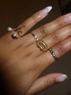 Mixade ringar i guld och silver - Säljer ett set med fyra ringar i olika stilar. En ring i guld med stjärndetalj och pärla, en tunn guldfärgad ring med liten rosa sten, en chunky guldfärgad ring formad som läppar och en silverfärgad ring med små kulor.💫✌🏼🤍