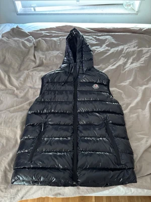 Svart dunväst med huva från Moncler - Snygg svart dunväst från Moncler med huva och dragkedja framtill. 