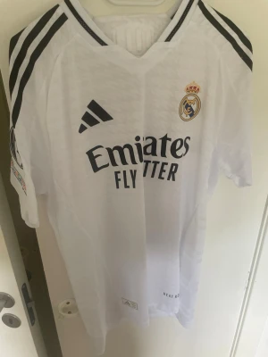 Real Madrid fotbollströja Adidas - Säljer en vit Real Madrid fotbollströja från Adidas med svarta detaljer och klubbmärke på bröstet. Tröjan har tryck med 'Mbappé' och nummer 9 på ryggen samt sponsorlogga på framsidan. Slim fit 