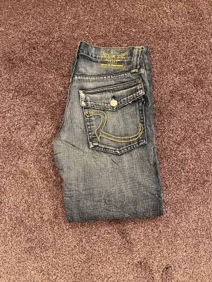 Rock and republic mörk blå slitna jeans - Säljer ett par mörk blå slitna jeans från Rock and republic med raka ben och slitningar. Jeansen har en stor gul broderad logga och fickor som har knappar. Jeansen har klassisk femficksmodell och kontrastsömmar. Perfekta för en avslappnad streetstil.
