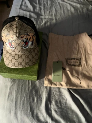 Gucci keps med tigertryck - Snygg keps från Gucci med klassiskt GG-mönster i beige och brunt samt två stora tigerhuvuden framtill. Baksidan är i svart mesh för extra ventilation. Kepsen kommer med originalpåse och tagg. Perfekt för dig som vill sticka ut med lyxig streetstyle.