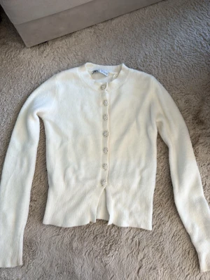 Vit kofta med pärlknappar från Zara - Supersöt vit kofta från Zara med långa ärmar och ribbade muddar. Koftan har dekorativa pärlknappar i blomform längs hela framsidan och är gjord i ett mjukt, stickat material. 