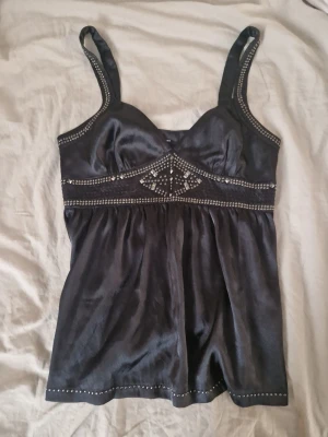 Svart satinlinnetopp från Guess - Supersnygg svart linnetopp från Guess i glansigt satinmaterial. Toppen har breda axelband och är dekorerad med silverfärgade nitar och små pärlor längs kanterna och under bysten. Upplever att storleken är stor för någon som brukar ha S-M