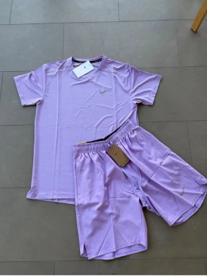 Ljuslila träningsset från Nike - Säljer ett ljuslila träningsset från Nike med matchande t-shirt och shorts. Setet är gjort i ett lätt och snabbtorkande material, perfekt för träning eller löpning. Båda plaggen har Nike-logga och shortsen har resår i midjan för skön passform.