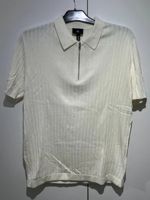 Vit polotopp med dragkedja från H&M - Klassisk vit polotopp från H&M med korta ärmar och diskret ribbat mönster. Toppen har en krage och en snygg dragkedja framtill som detalj. Perfekt till jeans eller kjol för en clean look.