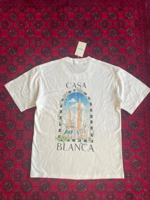 Vit Casa Blanca t-shirt med tryck - Säljer en vit t-shirt från Casa Blanca med ett stort färgglatt tryck på ryggen som föreställer en arkitektonisk scen med kupoler och palmer. T-shirten har klassisk passform och korta ärmar. Perfekt för dig som gillar unika prints och streetwear vibes.