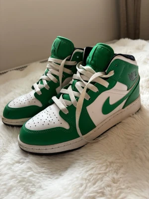 Nike Air Jordan 1 Mid grön/vit - Säljer ett par Nike Air Jordan 1 Mid sneakers i grön och vit färg. Skorna har klassisk Jordan-logga på sidan, gröna detaljer och vit bas i läder. Swoosh-loggan är grön och sulan är vit med svart yttersula. Perfekta för dig som gillar streetwear och snygga färgkombos. Se bilder för skick, fläck längst fram på skon men går säkert bort om man gör rent