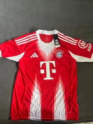 Bayern München matchtröja Adidas S - Röd och vit Bayern München matchtröja från Adidas i slim fit-modell. Tröjan har klubbmärke, Adidas-logga och sponsortryck framtill samt vita ränder på axlarna. Tillverkad i lätt och ventilerande material, perfekt för fotboll eller träning.