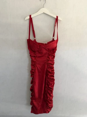 Röd satin klänning med rynk - Snygg röd klänning i satin med justerbara smala axelband och rynkade detaljer längs sidorna. Figurnära passform och axelbandslös look. Perfekt för dig som vill sticka ut med en elegant och trendig stil.