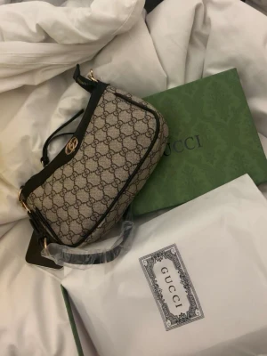 Gucci beige monogram handväska - Säljer en snygg beige handväska från Gucci med klassiskt GG-monogrammönster och svarta läderdetaljer. Väskan har guldfärgade metalldetaljer och justerbar axelrem. Perfekt för dig som vill ha en ikonisk och trendig accessoar. Box kommer med❤️