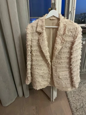 Beige fransig kavaj med struktur - Säljer en beige kavaj med cool fransig struktur och markerade slag. Kavajen har en unik design med horisontella rader av fransar över hela plagget och är helfodrad. Storlek XS, men mer en S.