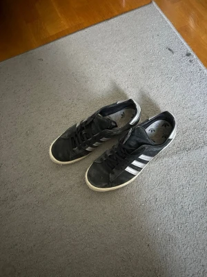Svarta Adidas sneakers med vita ränder - Klassiska svarta Adidas sneakers i mocka med de ikoniska tre vita ränderna på sidan och vit sula. Lågt skaft och snörning framtill. Perfekta för en avslappnad streetwear-look.