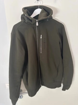 Grön hoodie från Sail Racing - Mörkgrön hoodie från Sail Racing med sned dragkedja och logga på bröstet. Tröjan har en stor huva med dragsko och ribbade muddar vid ärmslut och nederkant. Tillverkad i mjukt bomullsmaterial för skön känsla.